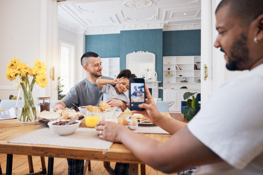 Two young fathers sit for breakfast with their toddler in a bright dining room. Sunlight streams in as the child poses for a photo, taken by one of her fathers. 