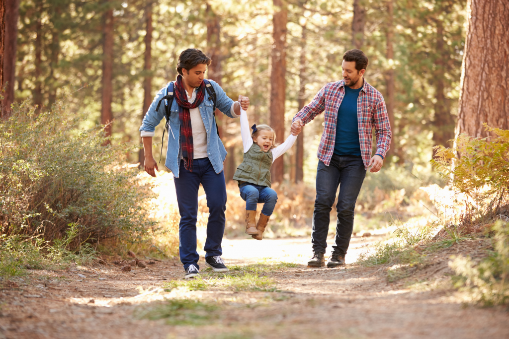Two fathers walk hand in hand, swinging their daughter between them as they laugh together in the forest. This scene captures authenticity, diversity, and the joy of shared family moments, illustrating &ldquo;family in motion,&rdquo; not just posed inclusion.