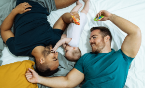 A joyful LGBTQIA+ family moment captured: two dads lounging on a bed with their content infant.