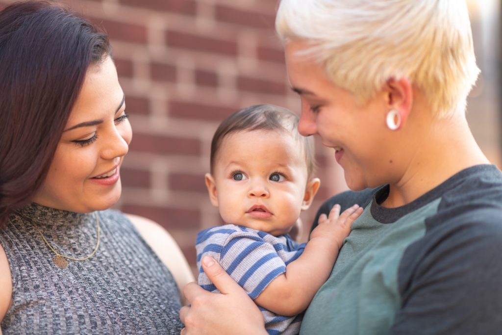 Two mums smile whilst holding their baby.