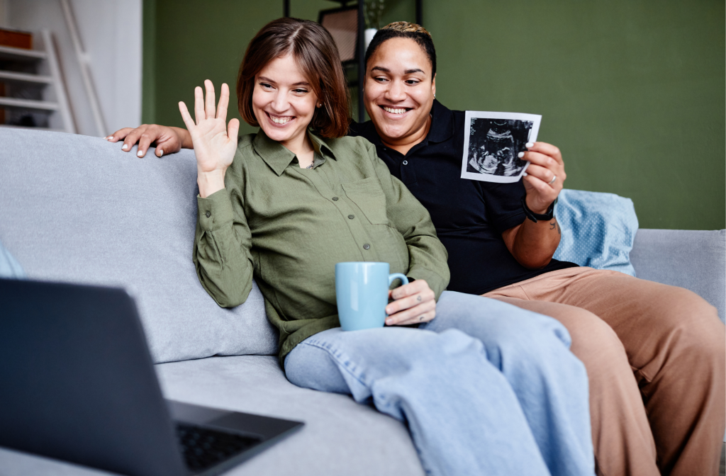 Two women on a sofa holding an ultrasound scan while speaking on a video call